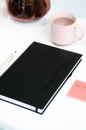 A black medical planner, white pen, pink sticky note, and pink mug sit on a white table