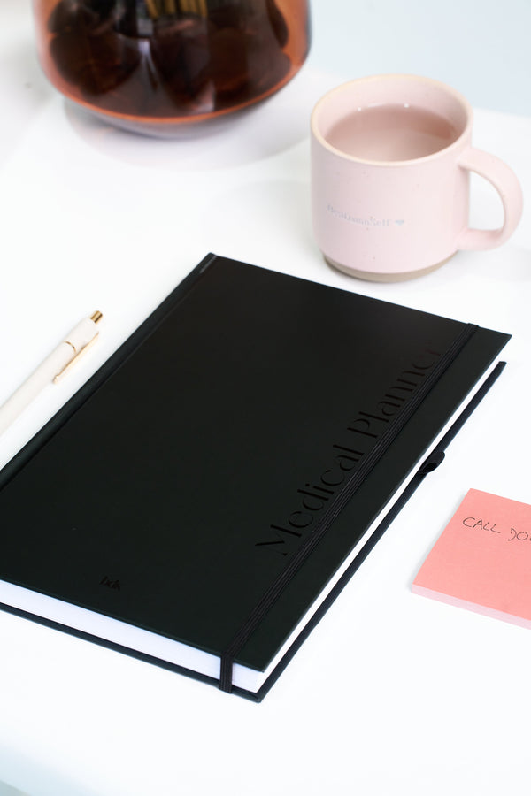 A black medical planner, white pen, pink sticky note, and pink mug sit on a white table