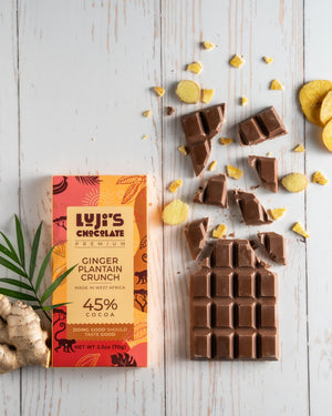 An unwrapped Luji's Ginger Plantain Crunch Chocolate bar broken into pieces alongside crispy plantain chips and fresh ginger, illustrating the bar's flavor components on a rustic white wooden background.