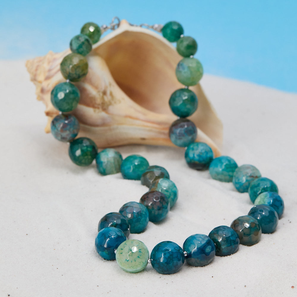 Necklace with blue and green beads on a white surface with a shell in the background